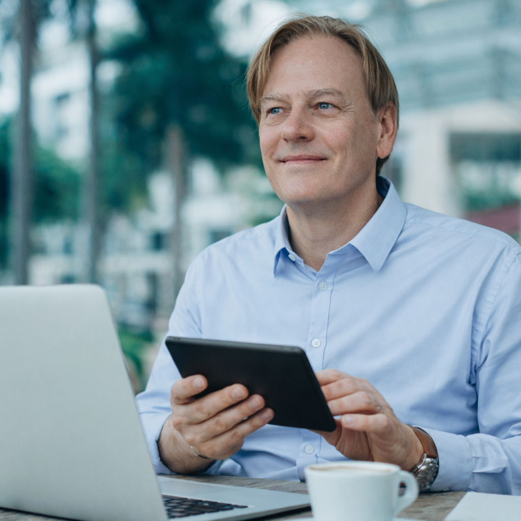 Homme avec une tablette, souriant, assis à une table à l'extérieur avec un ordinateur portable.